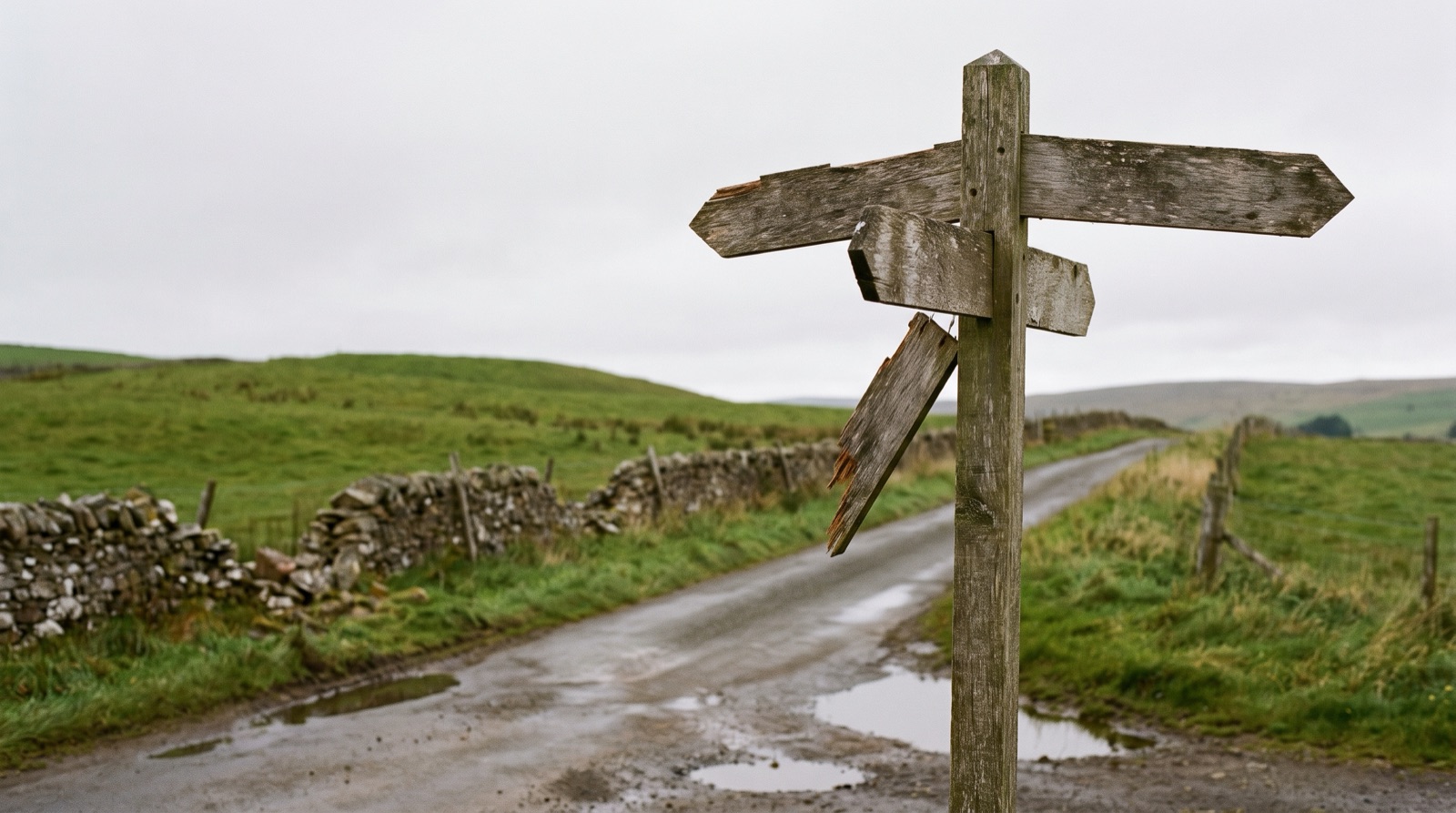 A weathered wooden signpost at a rural Scottish crossroads. One arm points on, one is askew and one hangs broken from the post.