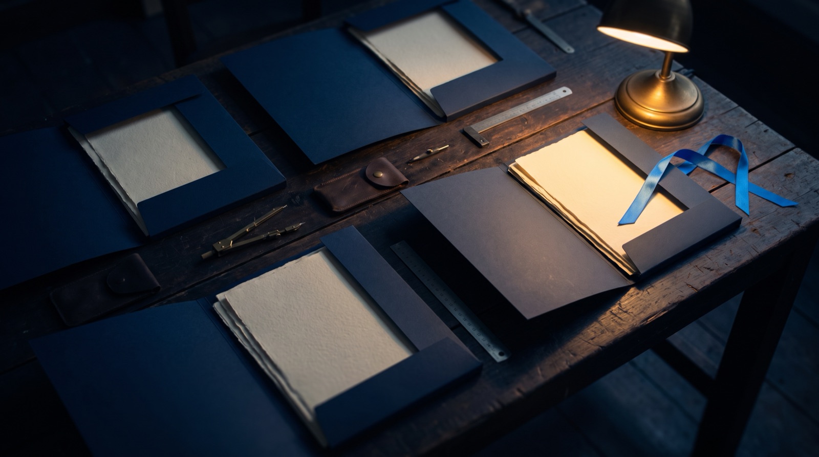 Navy blue folders laid open on a dark wooden workbench under a brass lamp, each holding sheets of heavy paper, with drawing instruments and a blue ribbon alongside.